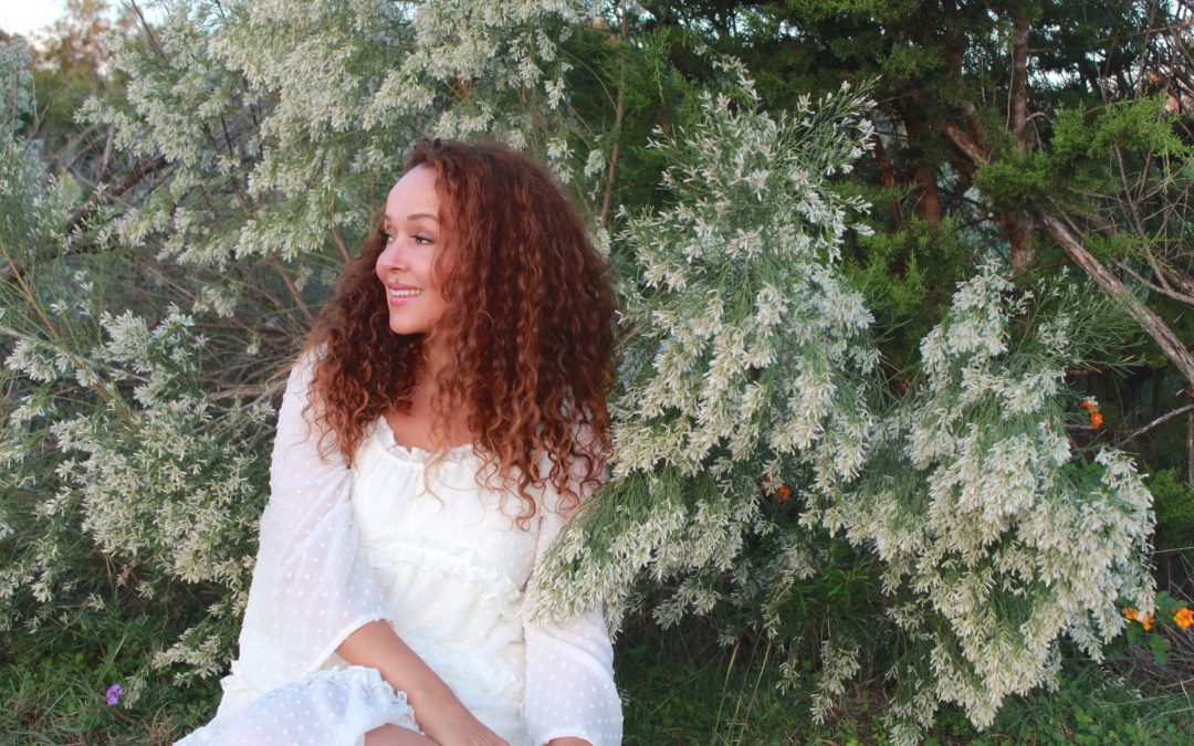 Author coach and podcast host Cynthia Occelli sitting on the ground in a white dress in front of trees and plants